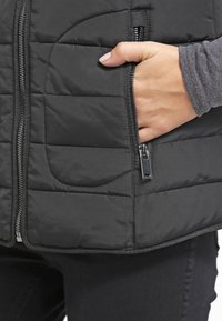 Černá vypolstrovaná vesta s zipem vpředu a bočními kapsami. Materiál je hladký s prošívanou texturou. Zipová kovová část je stříbrná.