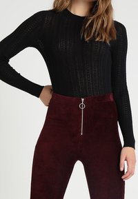 Haut en maille côtelée noire à manches longues associé à un pantalon en velours côtelé bordeaux taille haute avec une fermeture éclair devant et un détail de tirette circulaire.