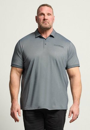 Uomo dalla corporatura muscolosa che indossa una polo grigia a maniche corte e pantaloni neri, in piedi davanti a uno sfondo chiaro uniforme.