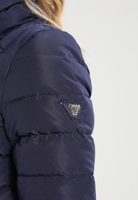 Marinblå vadderad jacka med quiltad textur, med en triangulär dekoration med glittrande detaljer på vänster axel.