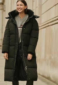 Cabană puf neagră cu un design lung, având un fermoar frontal și un guler supradimensionat. Asortată cu un pulover gri dedesubt.