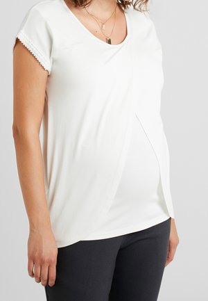 Mujer embarazada vistiendo una camiseta blanca de manga corta para maternidad con detalle frontal en capas y pantalones negros sobre un fondo liso.