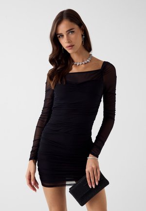 Schwarzes Minikleid mit transparenten langen Ärmeln, Raffung und quadratischem Ausschnitt. Accessoires: eine schwarze verzierten Clutch und silberne Schmuckstücke.