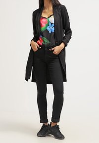 Americana negra larga sobre una camiseta de flores colorida, combinada con jeans negros ajustados y zapatillas negras. Tejido suave, diseño casual.