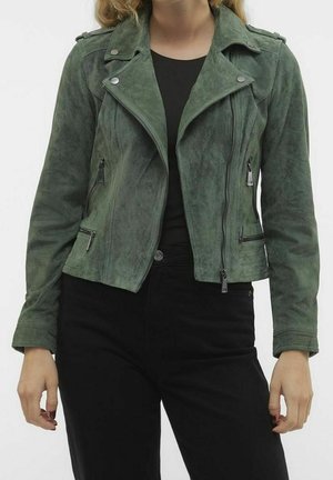 Personne portant une veste de motard en daim verte avec une fermeture éclair asymétrique sur une chemise noire et un pantalon noir, debout devant un fond uni.
