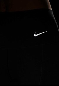 Czarne legginsy sportowe o gładkiej fakturze, z białym logo Nike w kształcie łuku na pasie. Brak innych wzorów lub akcentów.