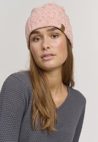 Lys rosa strikket lue med ribbestruktur og et lite brunt logo-merke. Stoffet ser mykt og teksturert ut.