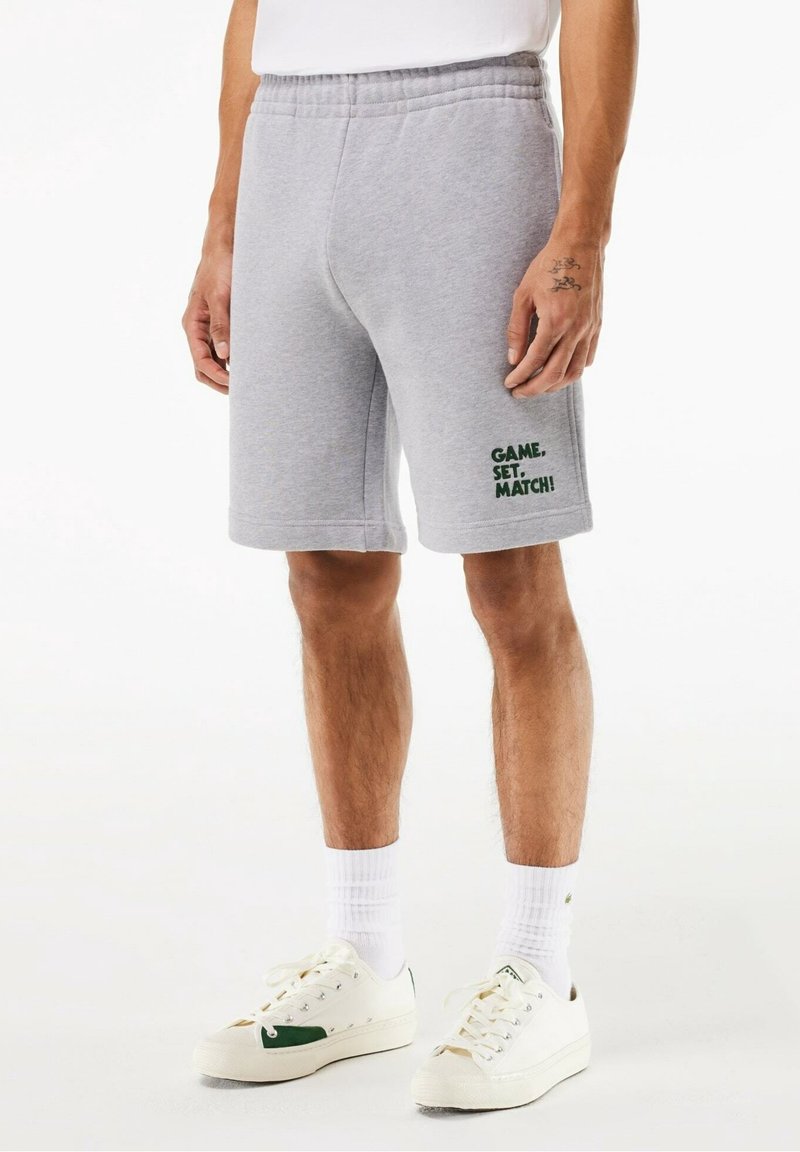 Shorts en coton gris avec une taille élastique et des poches latérales. Présente le texte vert "GAME, SET, MATCH!" sur la jambe gauche.