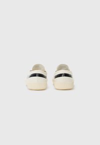 Beige Canvas-Slipper mit einem schwarzen Akzentstreifen und einer strukturierten Gummisohle, die ein minimalistisches Design und eine abgerundete Form aufweisen.