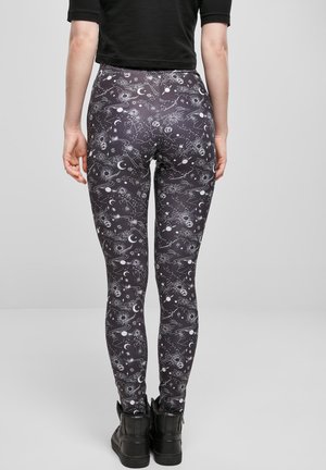 Juodos leggingai su dangaus tema, kuriuose pavaizduoti mėnuliai, žvaigždės ir planetos baltos spalvos. Medžiaga turi glotnų, tamprų tekstūrą.