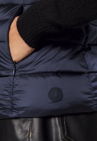 Marineblaue Steppweste mit glatter Textur und elastischen schwarzen Strickbündchen. Verfügt über eine Reißverschlusstasche und ein kreisförmiges Logopatch auf der Vorderseite.