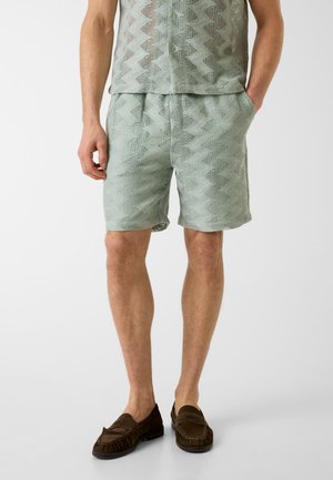 Mężczyzna ubrany w jasnozielone szorty zygzakowate wzory i pasującą koszulę, w ciemnobrązowych mokasynach, stojący na jednolitym tle.