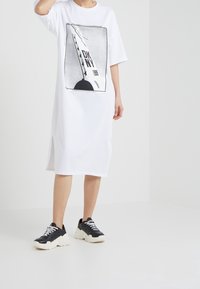 Biała sukienka t-shirtowa z czarnym nadrukiem graficznym, okrągłym dekoltem i krótkimi rękawami z bocznymi rozcięciami. Połączona z czarno-białymi sneakersami.