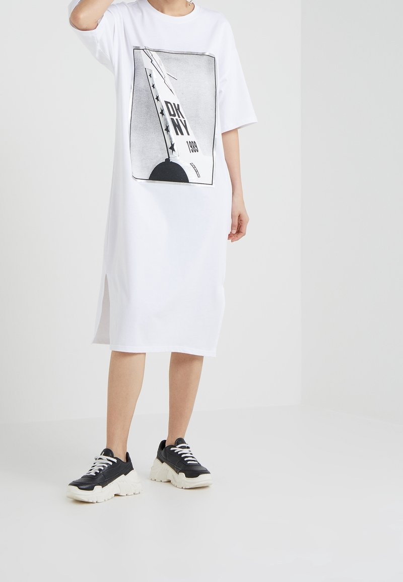 Biała sukienka t-shirtowa z czarnym nadrukiem graficznym, okrągłym dekoltem i krótkimi rękawami z bocznymi rozcięciami. Połączona z czarno-białymi sneakersami.