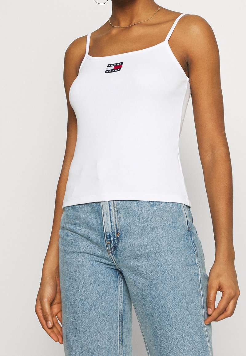 Femme portant un débardeur blanc à fines bretelles avec le logo Tommy Jeans et un jean taille haute bleu clair sur fond blanc.