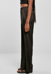Vue de côté d'une personne portant un pantalon large en satin noir, un crop top noir et des sandales noires, debout devant un fond gris uni.