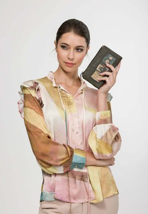 Frau mit dunklen Haaren in einer mehrfarbigen Bluse hält eine schwarze Geldbörse mit dekorativen Patches nahe an ihr Gesicht und blickt leicht zur Seite.