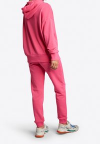 Różowa bluza z kapturem i dopasowane dresy wykonane z miękkiego materiału, charakteryzujące się luźnym krojem i kapturem. Sportowe buty w mieszanych kolorach i akcentach dopełniają całość.
