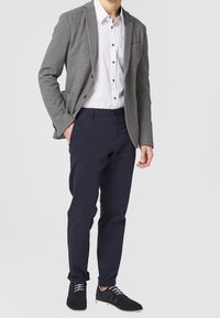 Blazer grigio con rever a punta e due tasche frontali, camicia bianca con bottoni, pantaloni blu scuro e scarpe nere con lacci bianchi.