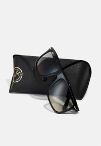 Ochelari de soare negri Ray-Ban cu lentile degradate, așezați într-un unghi pe un etui texturat din piele neagră, având un logo auriu.