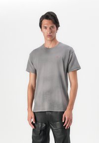 T-shirt de algodão cinza com mangas curtas, gola redonda e uma textura subtil. Apresenta um pequeno logo vermelho no peito. Usado com calças de couro pretas.