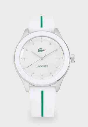 Montre en silicone blanche avec boîtier en acier inoxydable argenté, accents verts, et un cadran minimaliste avec des index en cristal et un logo Lacoste.
