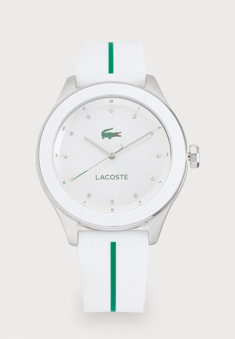 Baltas silikono laikrodis su sidabriniu korpusu, žaliais akcentais ir minimalistišku ciferblatu, kuriame yra kristaliniai valandų žymenys ir Lacoste logotipas.