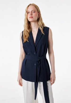 Donna bionda che indossa un blazer senza maniche a righe blu navy con cintura e pantaloni bianchi, in piedi davanti a uno sfondo bianco semplice.