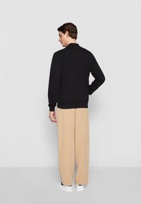 Svart genser med høy hals, ribbestrikkede ermer og nederkant. Kombinert med beige vide bukser og hvite joggesko, sett fra baksiden.