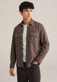 Ruda languota marškinėliai su dviem krūtinės kišenėmis ir baltais mygtukais, dėvimi virš šviesiai pilkos marškinėlių, derinami su tamsiomis kelnėmis.