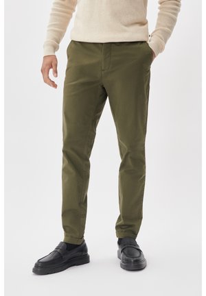 Mężczyzna ubrany w oliwkowozielone spodnie slim fit, beżowy sweter i czarne mokasyny, stojący z jedną ręką w kieszeni na białym tle.