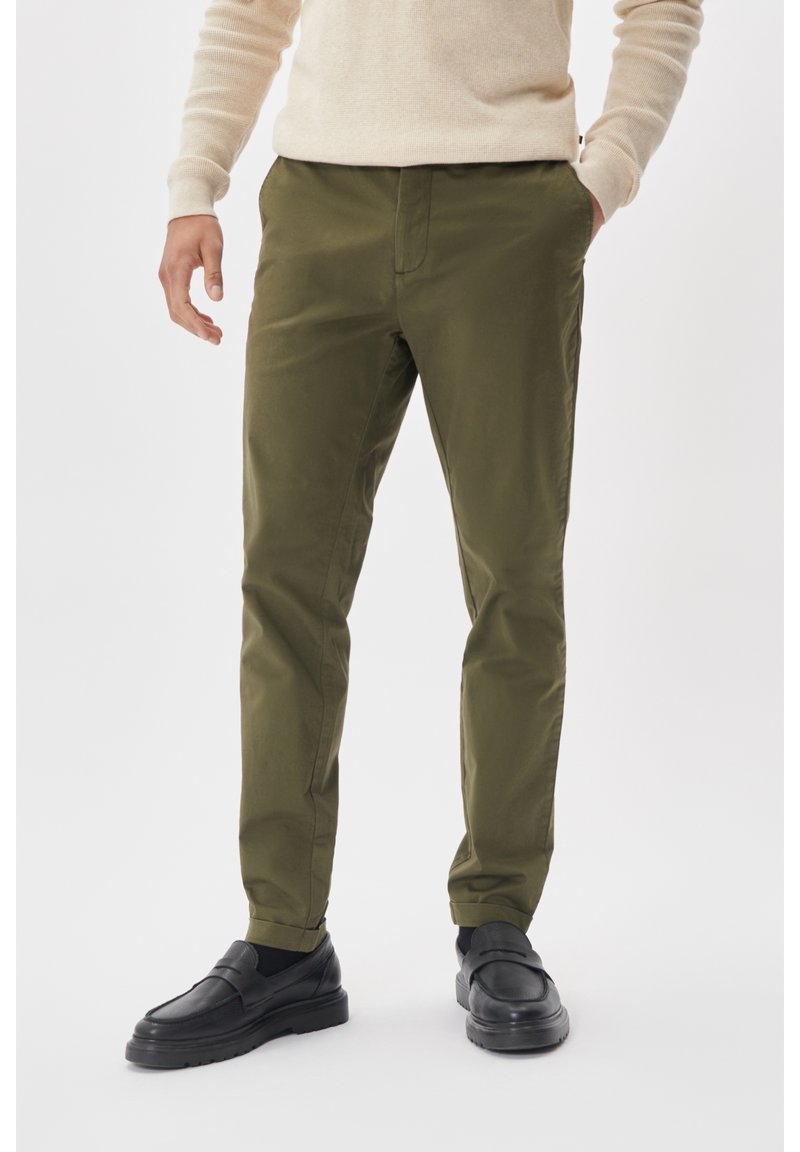 Homme portant un pantalon slim vert olive, un pull beige et des mocassins noirs, debout avec une main dans la poche sur fond blanc.