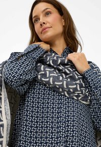 Vrouw die een gestreepte marineblauwe en witte sjaal aanpast, gekleed in een bijpassend gestreept marineblauw shirt, tegen een witte achtergrond.