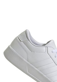 White sneaker with a textured synthetic upper, padded collar, and flat white laces. Features a grey rubber sole and subtle side stripes.