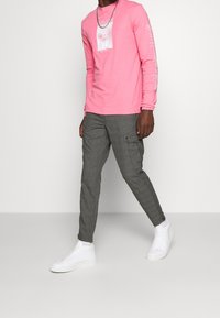 Růžová košile s dlouhým rukávem a bílým grafickým vzorem, v kombinaci se šedými kostkovanými cargo kalhotami a bílými teniskami; má volný střih a dlouhé rukávy.