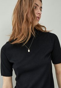 Woman with light brown hair wearing a black short-sleeve top and a thin chain necklace with a small rectangular pendant, looking sideways.