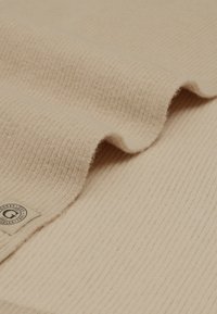Manta beige con textura suave y rayas, con una etiqueta tejida en una esquina. La tela presenta suaves ondas a lo largo de los bordes.