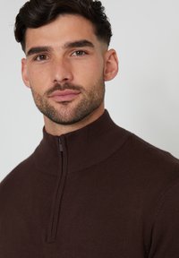 Maglione zip-neck marrone in un morbido materiale a maglia fine con colletto e orlo a costine. Presenta una chiusura a mezza zip nella parte anteriore.