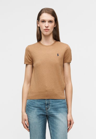 Haut en tricot à manches courtes marron avec un col rond, orné d'un petit logo bleu marine sur le côté gauche de la poitrine, assorti à un jean bleu clair.