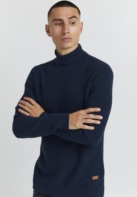 Mornarsko pulover s turtleneck ovratnikom, izdelan iz teksturirane pletenine, z dolgimi rokavi in majhnim rjavim logotipom na spodnji strani.
