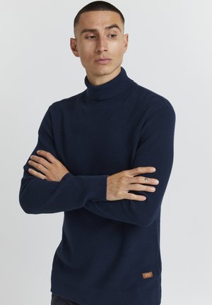 Granatowy sweter z golfem wykonany z teksturowanej dzianiny, z długimi rękawami i małą brązową naszywką z logo na dolnej stronie.