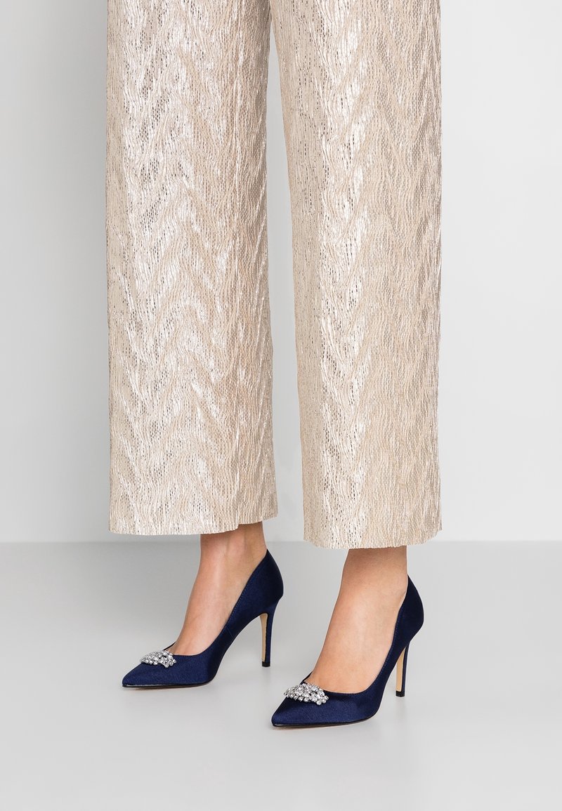 Personne portant un pantalon large texturé doré clair et des escarpins bleu marine pointus avec des ornements de bijoux sur les orteils.