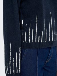 Pull bleu marine foncé avec des bandes verticales de sequins argentés sur la manche et l’ourlet, porté avec un jean bleu.