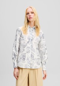 Camicia bianca con bottoni, caratterizzata da un motivo di schizzi architettonici blu, maniche lunghe e orlo arrotondato. Indossata con pantaloni larghi beige.