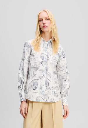 White button-up shirt featuring a blue architectural sketch pattern, long sleeves, and a rounded hem. Worn with beige wide-leg trousers.