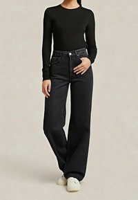 Femme portant un haut noir à manches longues, un jean noir taille haute à jambes larges, et des baskets blanches, debout devant un fond uni.