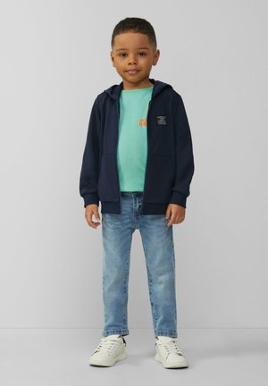 Marine rits-trui over een mintgroen shirt, gecombineerd met lichtblauwe jeans en witte sneakers. Opvallend logo op de borst en een casual pasvorm.