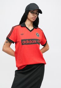 Czerwona koszulka w pionowe paski, czarny kołnierz i logo. Do tego czarna spódnica i czapka z logo Adidas w czerni.