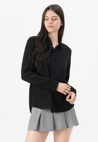 Zwarte langwerpige button-up shirt met een kraag, gemaakt van een gladde stof. Het shirt heeft een relaxte pasvorm en subtiele zijsplitten aan de zoom.