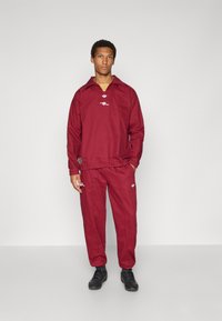 Μπορντό φόρμα από ελαφρύ ύφασμα, με γιακά, διακοσμητικά λογότυπα και ελαστικά μανσέτα στα παντελόνια. Φορεμένη με μαύρα αθλητικά παπούτσια.